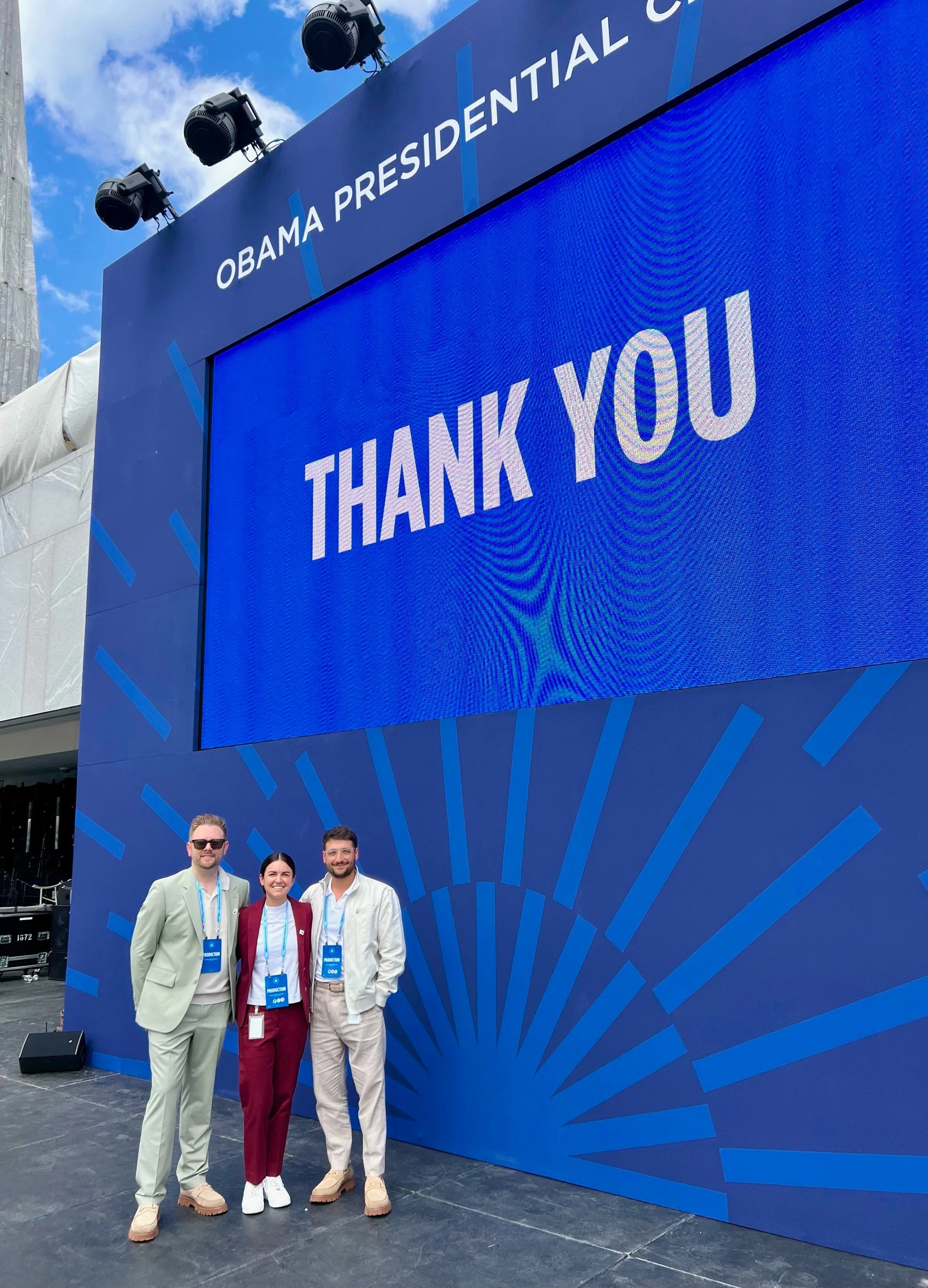 Three people standing in front of a large blue sign with the words 'Thank You' and 'Obama Presidential Center' at an outdoor event. The individuals are wearing conference badges and are dressed in business casual attire.