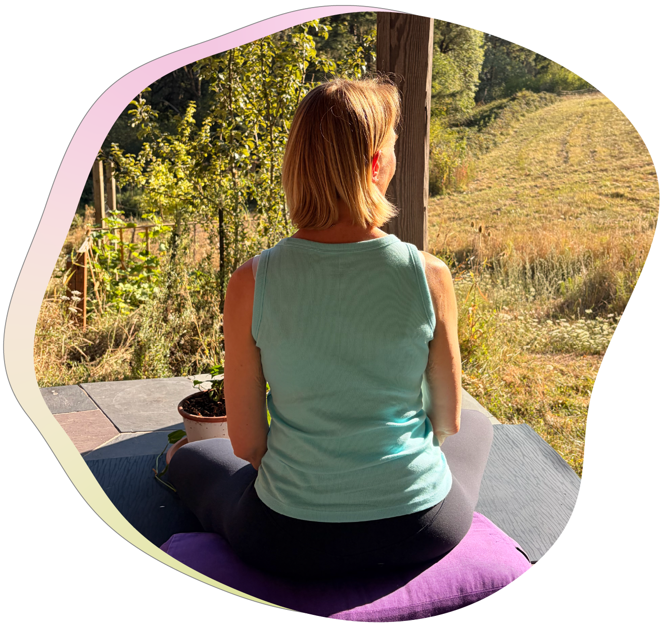 A woman with blond hair practicing yoga outdoors on a purple mat, sitting cross-legged and facing a scenic view of greenery and rolling hills.