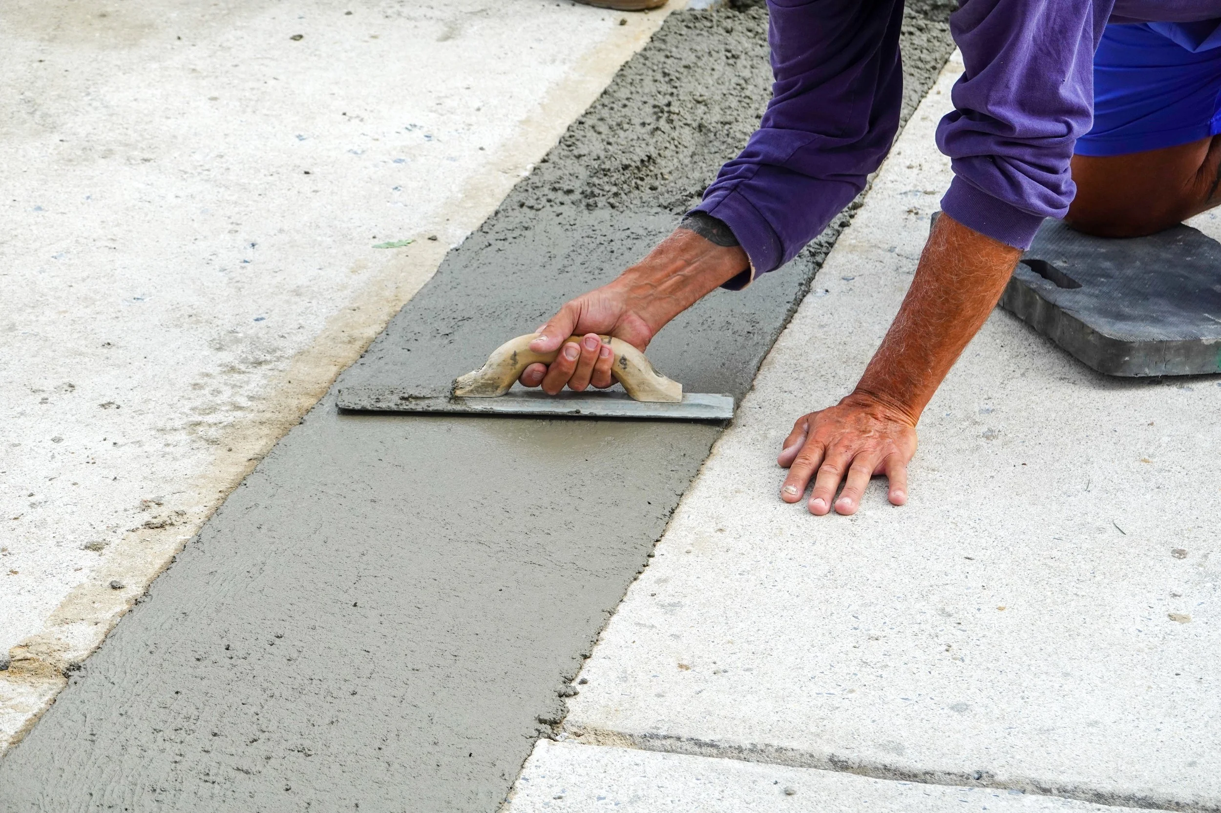 A person kneeling down on their knees while smoothing fresh cement on the ground with a trowel.