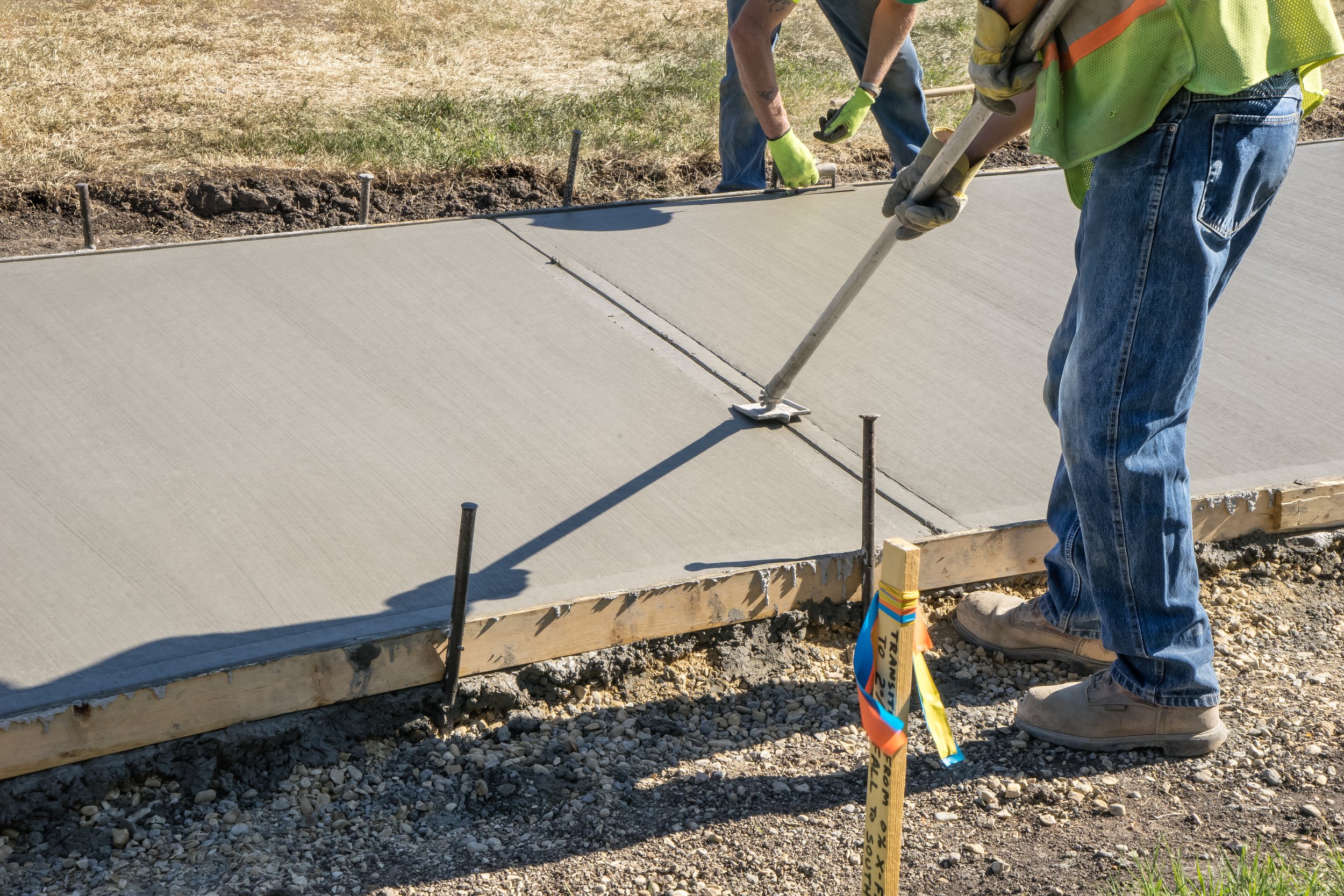 People laying down fresh concrete on a sidewalk with tools and safety gear visible.