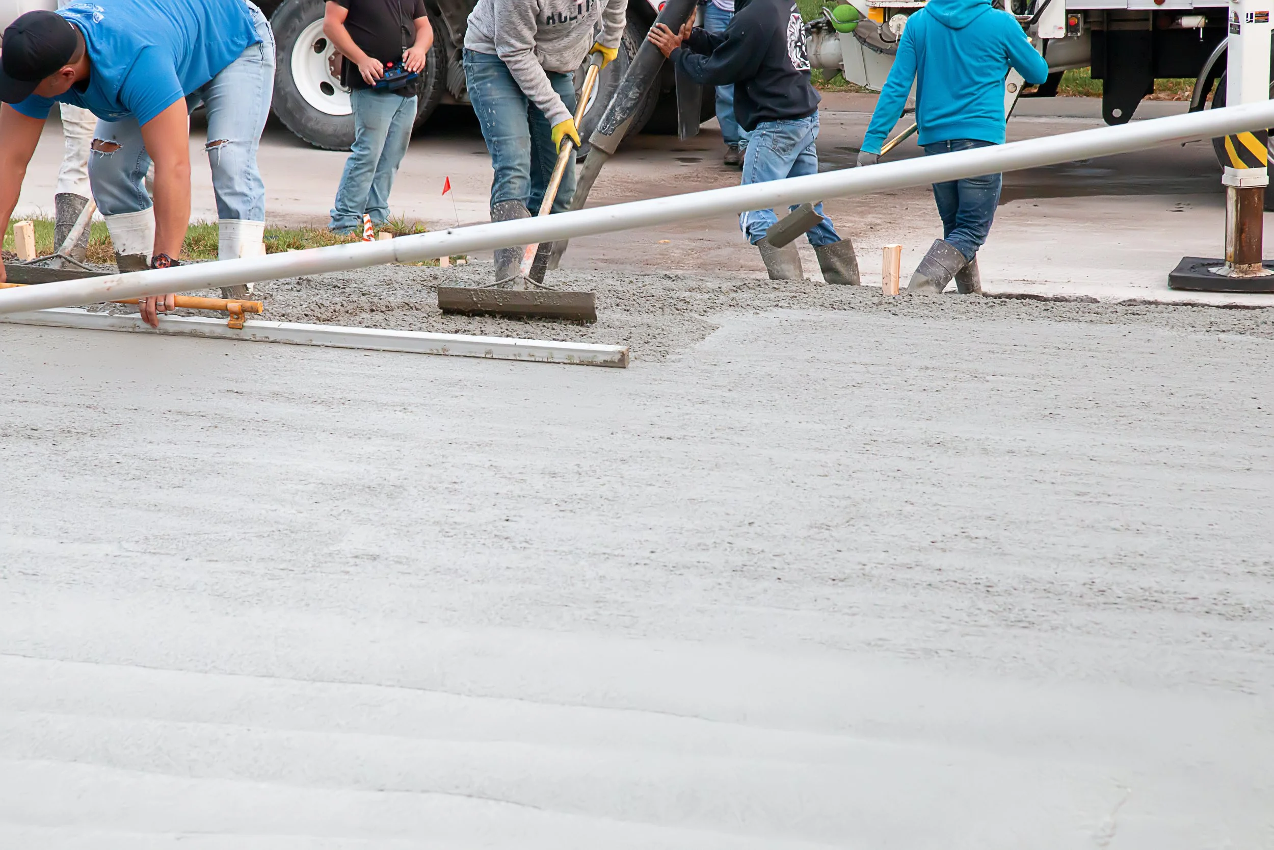 Construction workers pouring and leveling concrete on a sidewalk or road surface, with traffic barriers and machinery nearby.