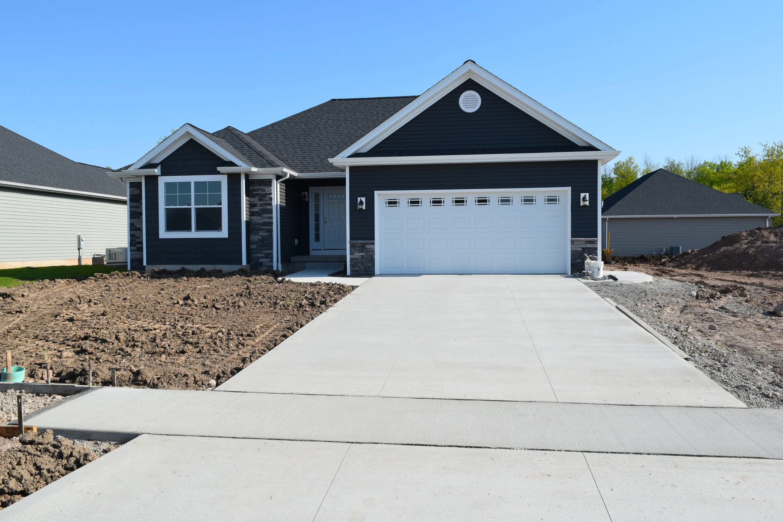 A newly constructed house with dark blue siding, white trim, and a white garage door, with a concrete driveway and dirt yard in the front yard, under a clear blue sky.