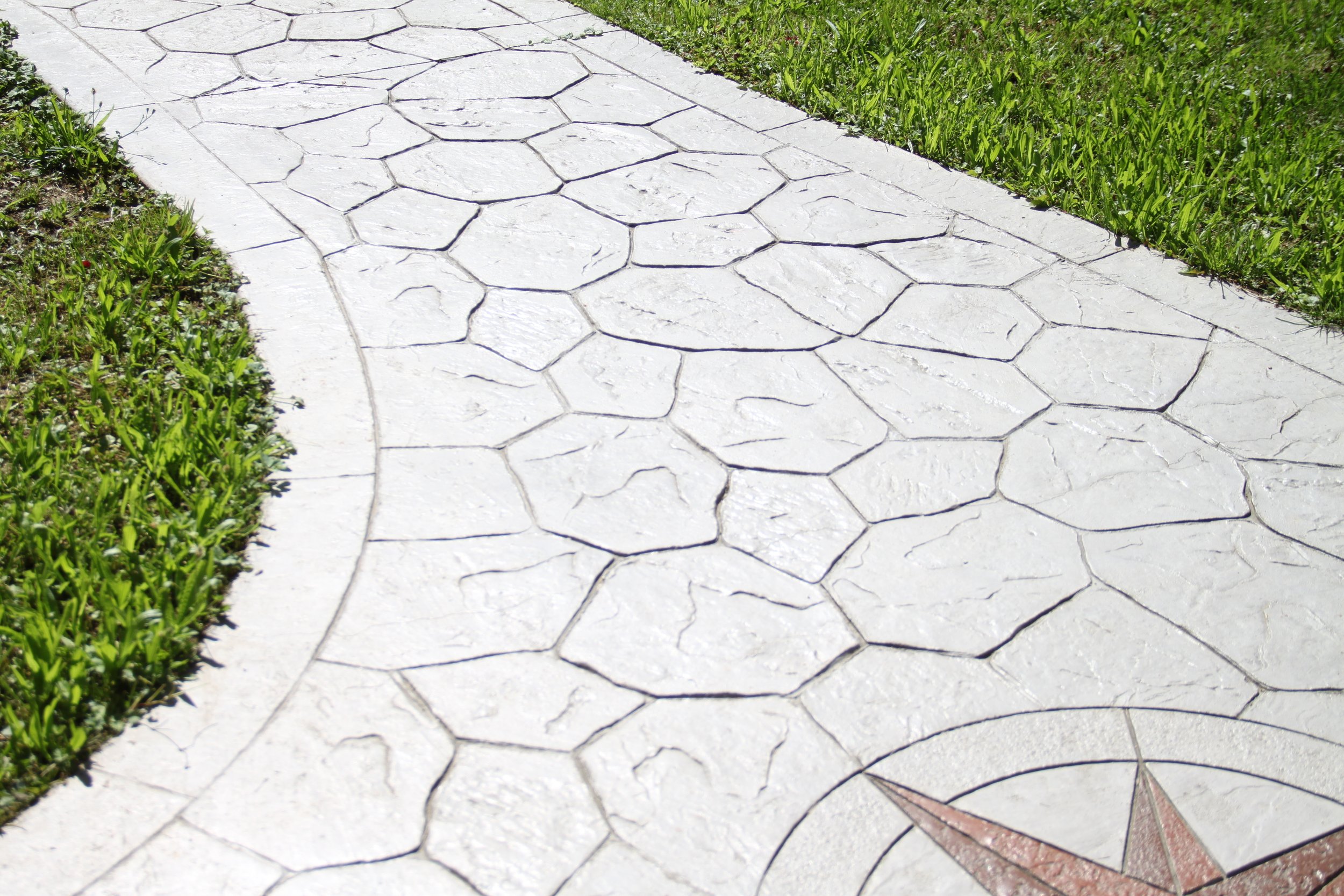 A concrete sidewalk with hexagonal and irregular stones, bordered by green grass with some weeds.