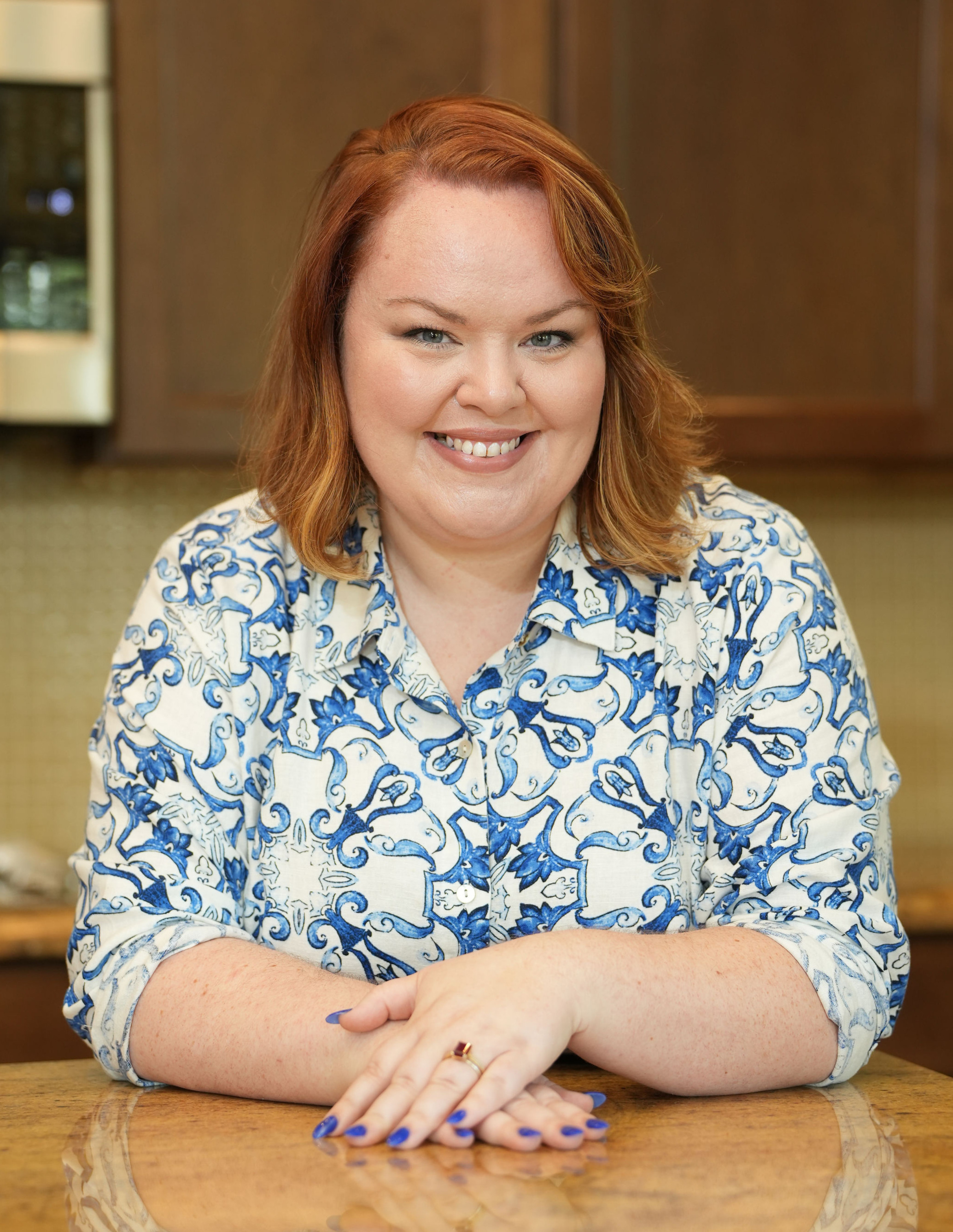 A woman with red hair, wearing a blue and white patterned shirt, sitting at a wooden table with her arms crossed, smiling at the camera.