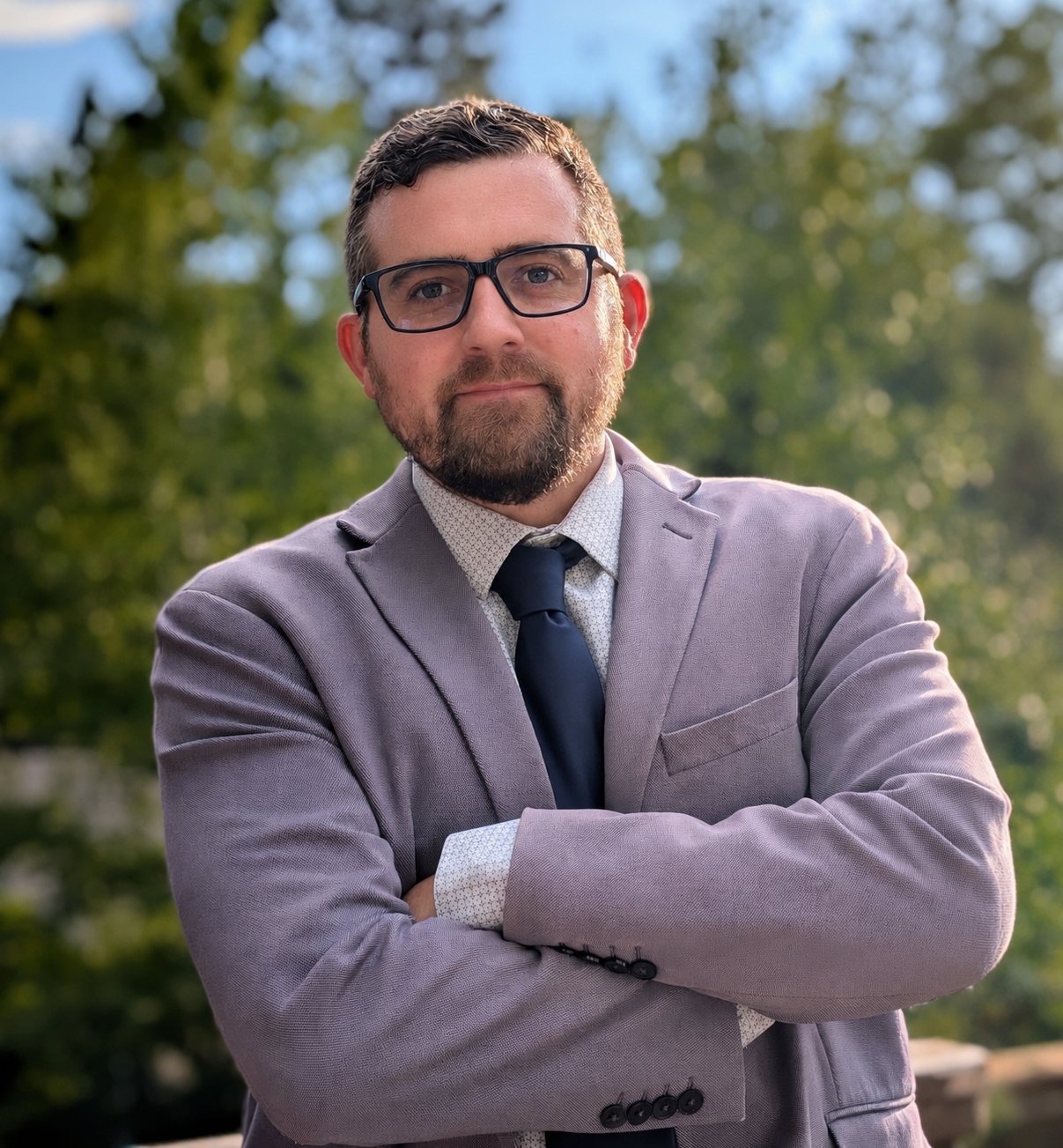 A man in a gray suit with a white shirt, dark blue tie, glasses, and a beard, standing outdoors with arms crossed, with blurred green trees in the background.
