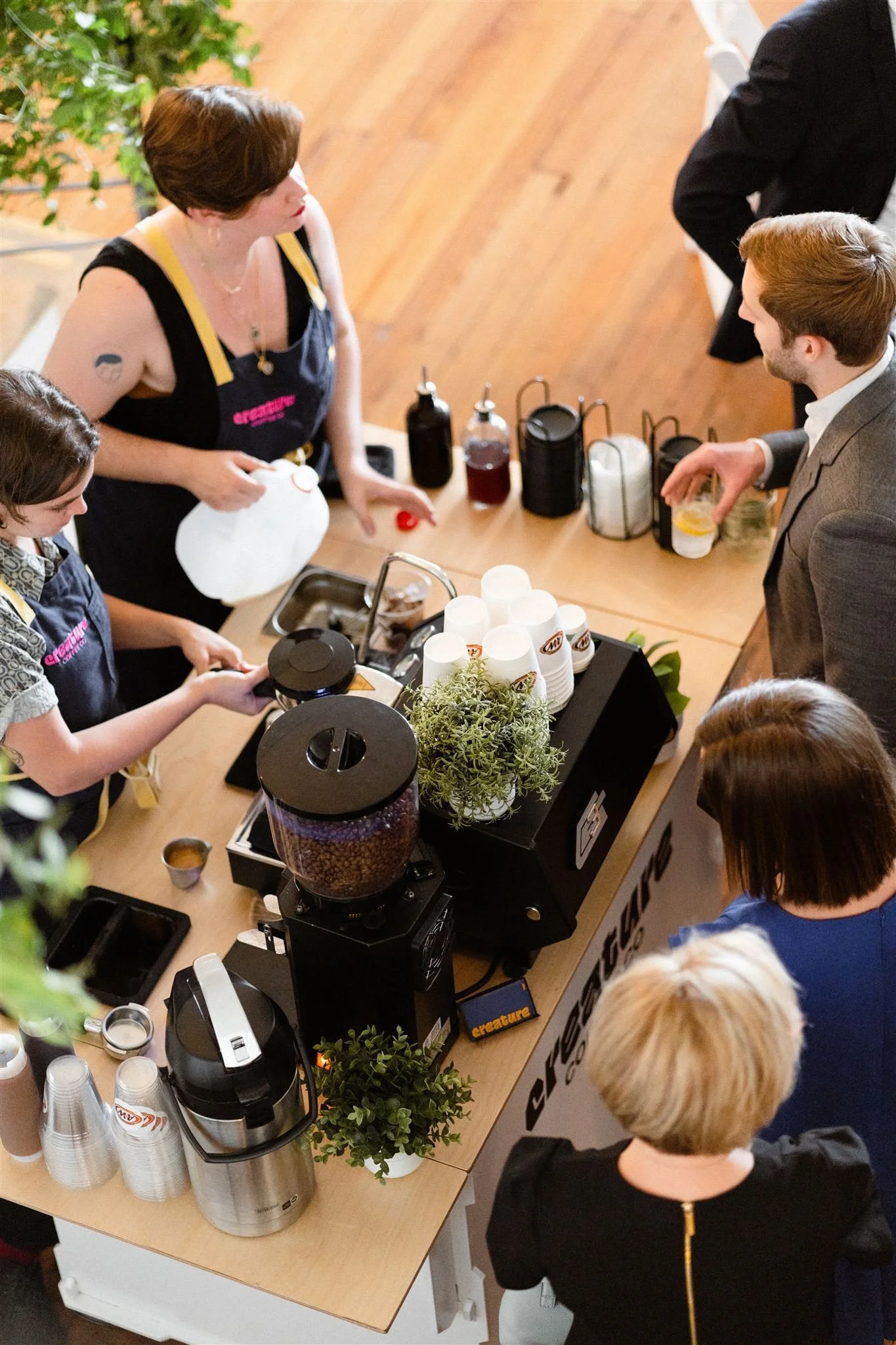 People gathered around a table with coffee, cups, and coffee-making equipment, engaging in a demonstration or conversation in a well-lit indoor setting.