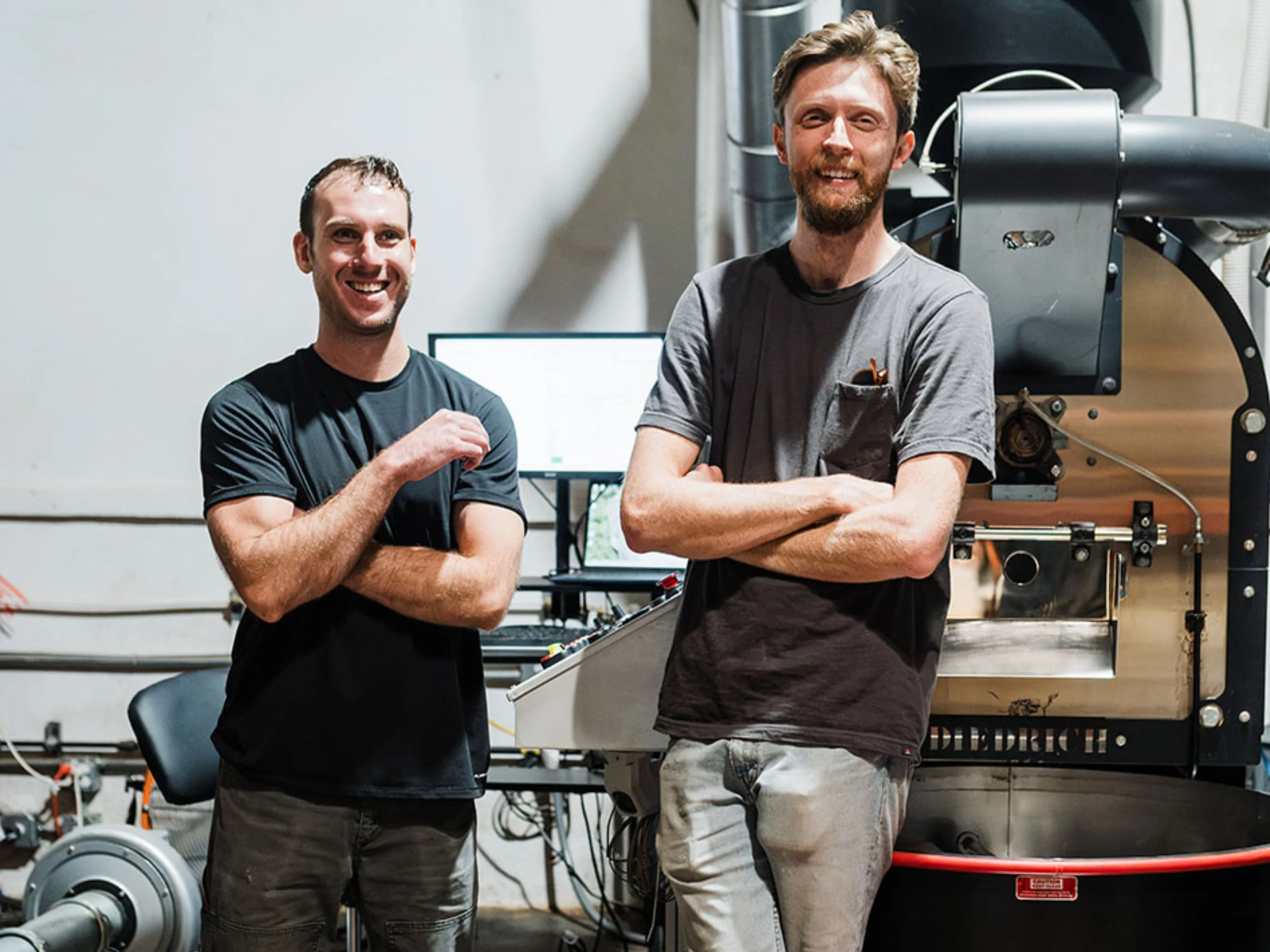 Two smiling young men with beards standing in a workshop, one with arms crossed, near machinery and a computer monitor.