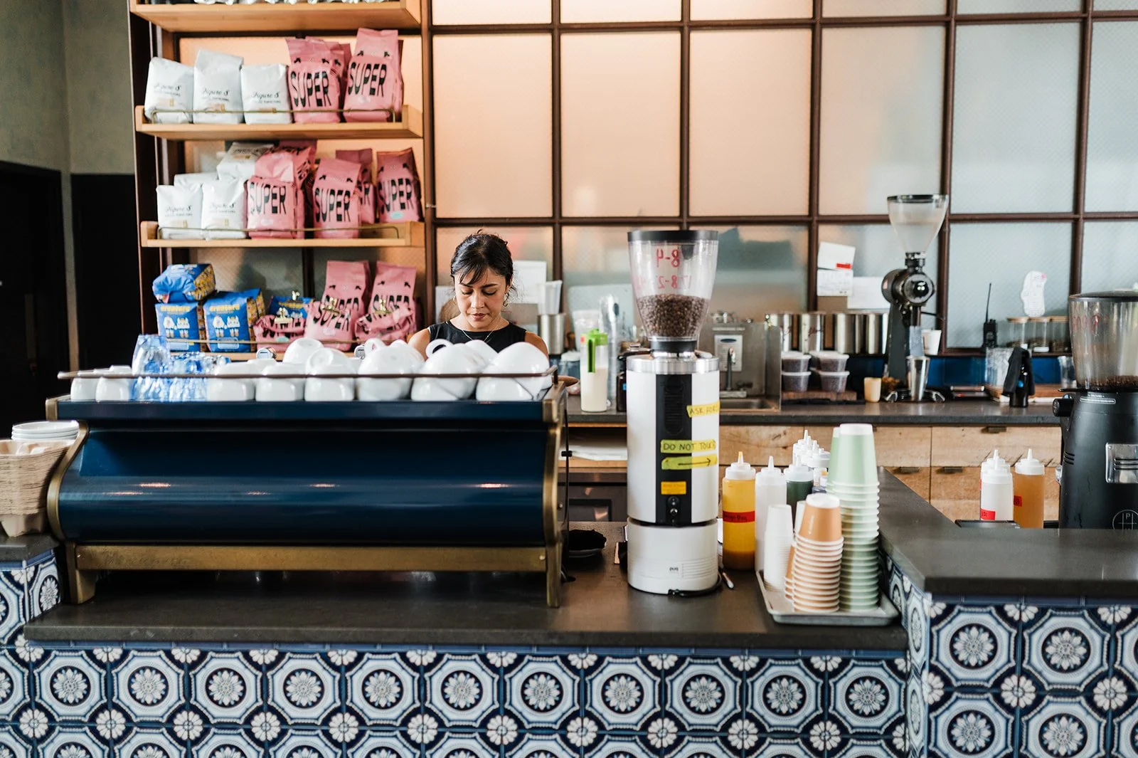A woman working behind a coffee counter with various coffee supplies, cups, and a large grinder, in a cafe with patterned tiles on the counter and shelves of tea or coffee packages in the background.