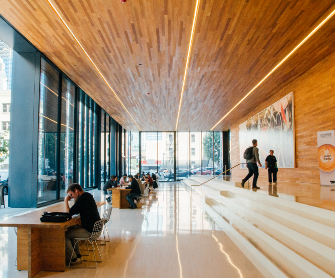 Modern lobby with large glass windows, wooden walls and ceiling, and seating area with people working and talking.