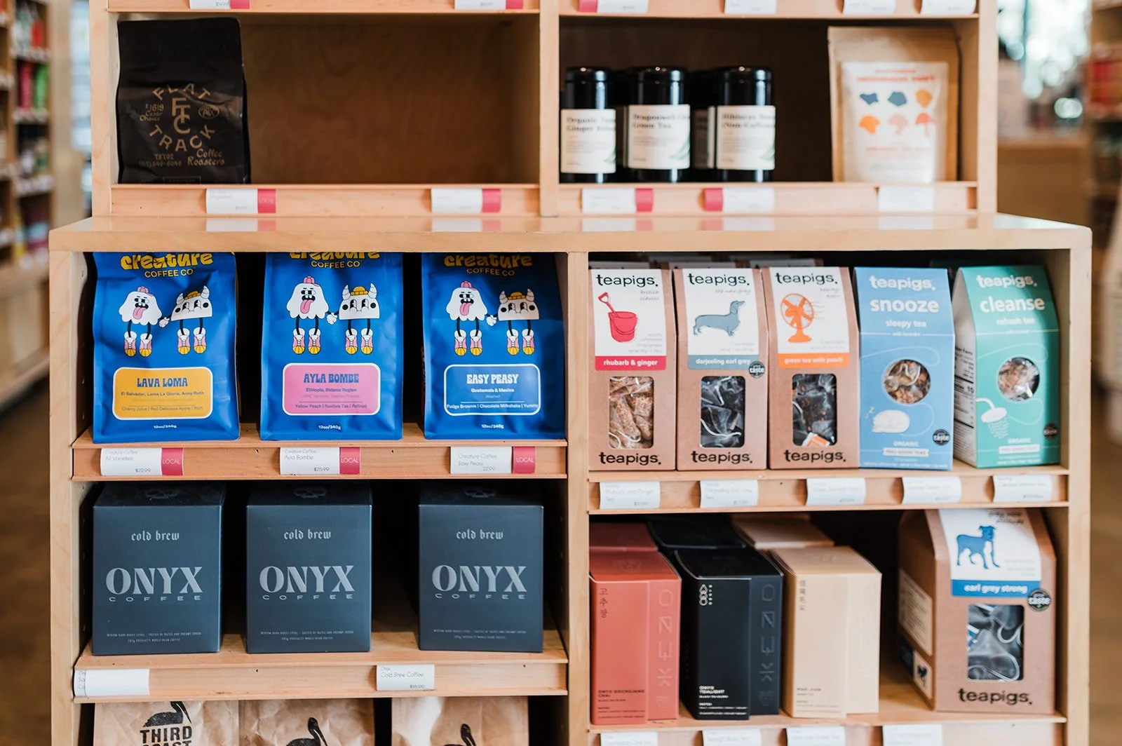Wooden store display shelf filled with various coffee products, including bags of Creature Coffee in blue packaging featuring cartoon dogs, boxes of Teapigs herbal teas, and Onyx Coffee in black packaging.