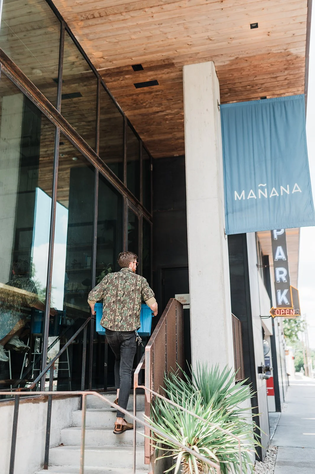 A man wearing a floral shirt and sunglasses carrying a blue box up the stairs of a building with large glass windows and a modern exterior. A blue banner with the word 'MAÑANA' hangs above, and a sign indicates the park is open.