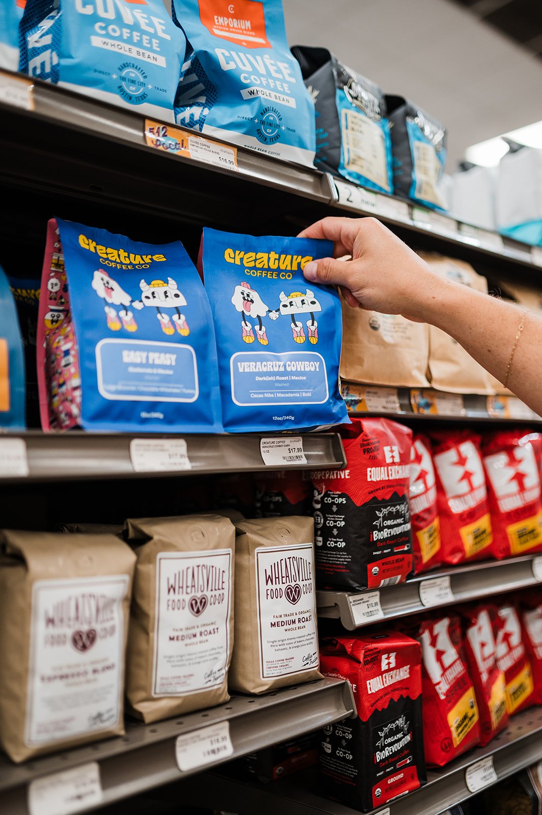 A person shopping for coffee at a grocery store, holding a bag of Creature Coffee Veracruz Cowboy coffee on a store shelf surrounded by other brands.