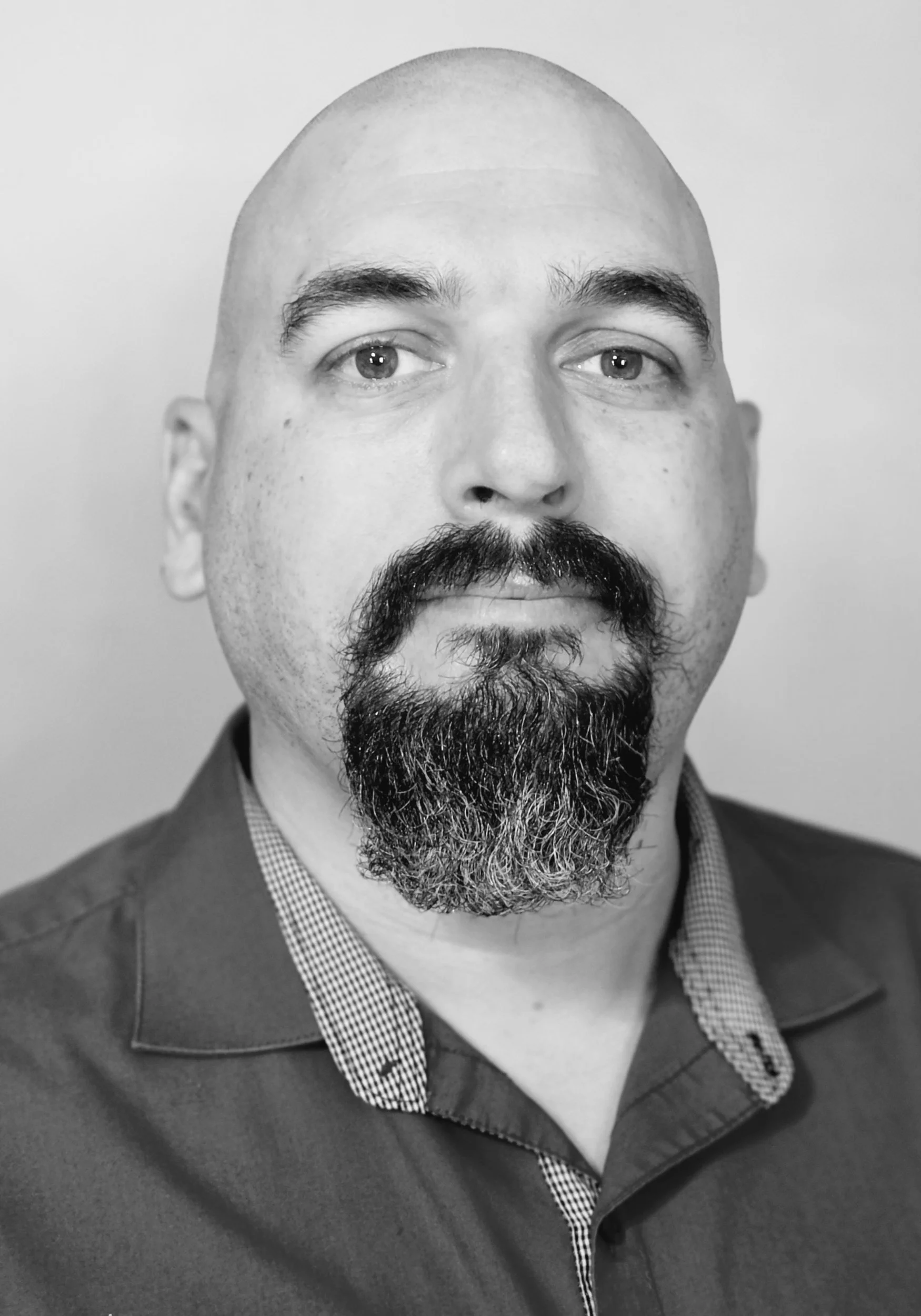 Black and white portrait of a middle-aged man with a bald head, thick eyebrows, and a full beard and mustache, wearing a collared shirt underneath a jacket.