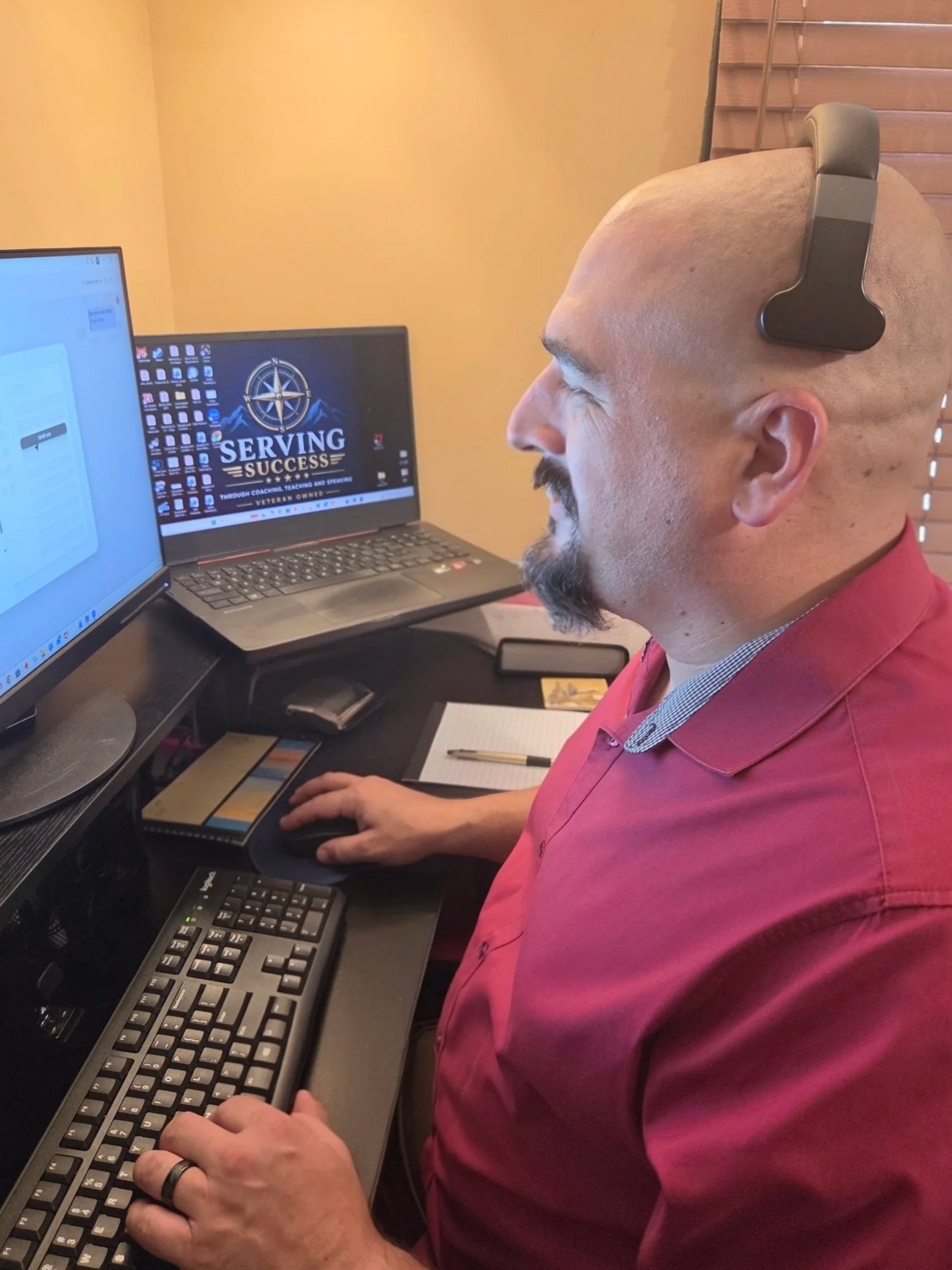 A man with a bald head and a beard wearing a maroon shirt, sitting at a desk with two computer monitors, typing on a keyboard, with a notebook and a pen on the desk.