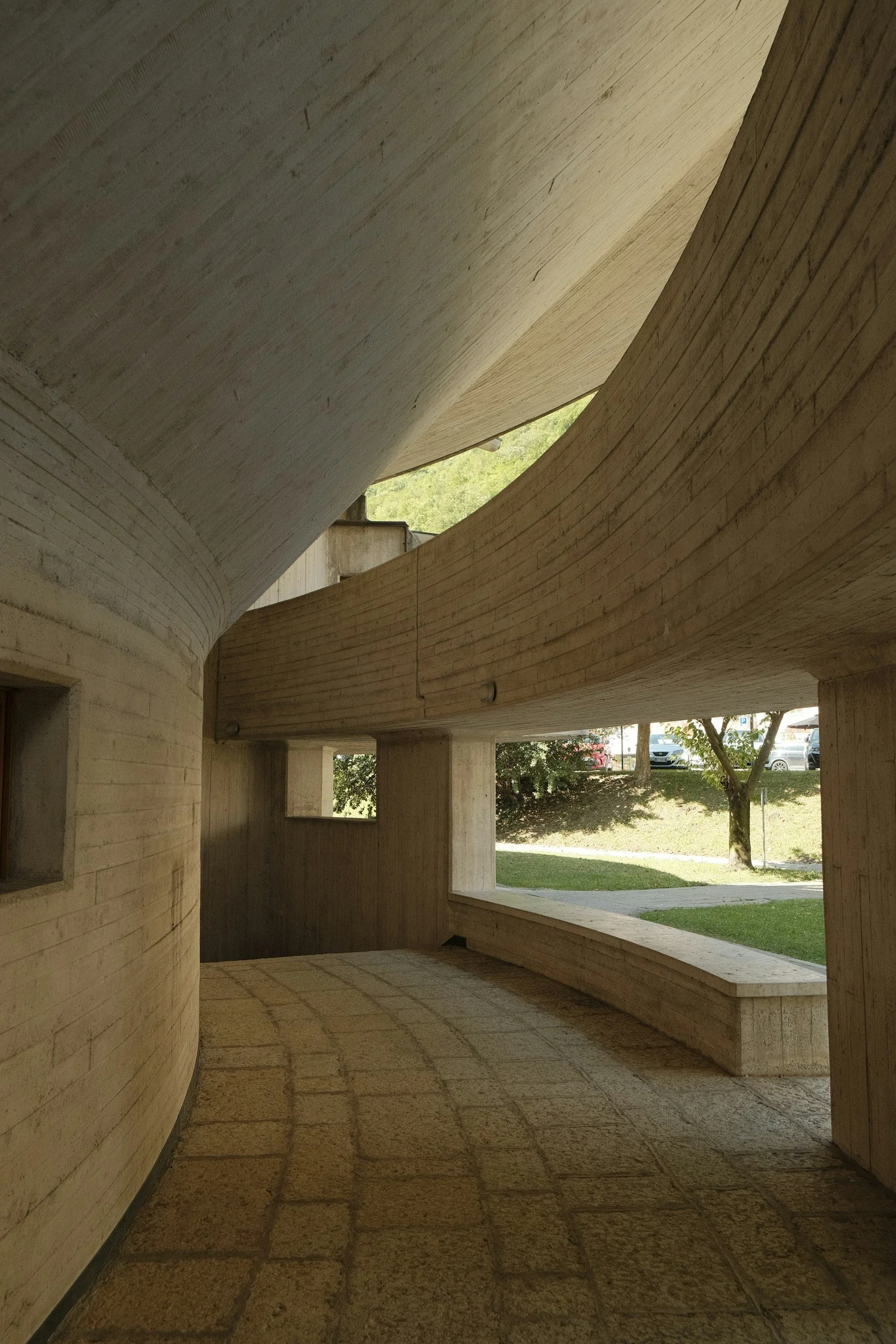 Vue intérieure d'une structure moderne en béton avec un plafond courbé et de grandes fenêtres laissant entrer la lumière naturelle, donnant sur un espace extérieur avec des arbres et une route.