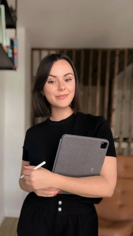 Une femme souriante tenant une tablette et un stylet, portant un t-shirt noir dans un intérieur moderne.