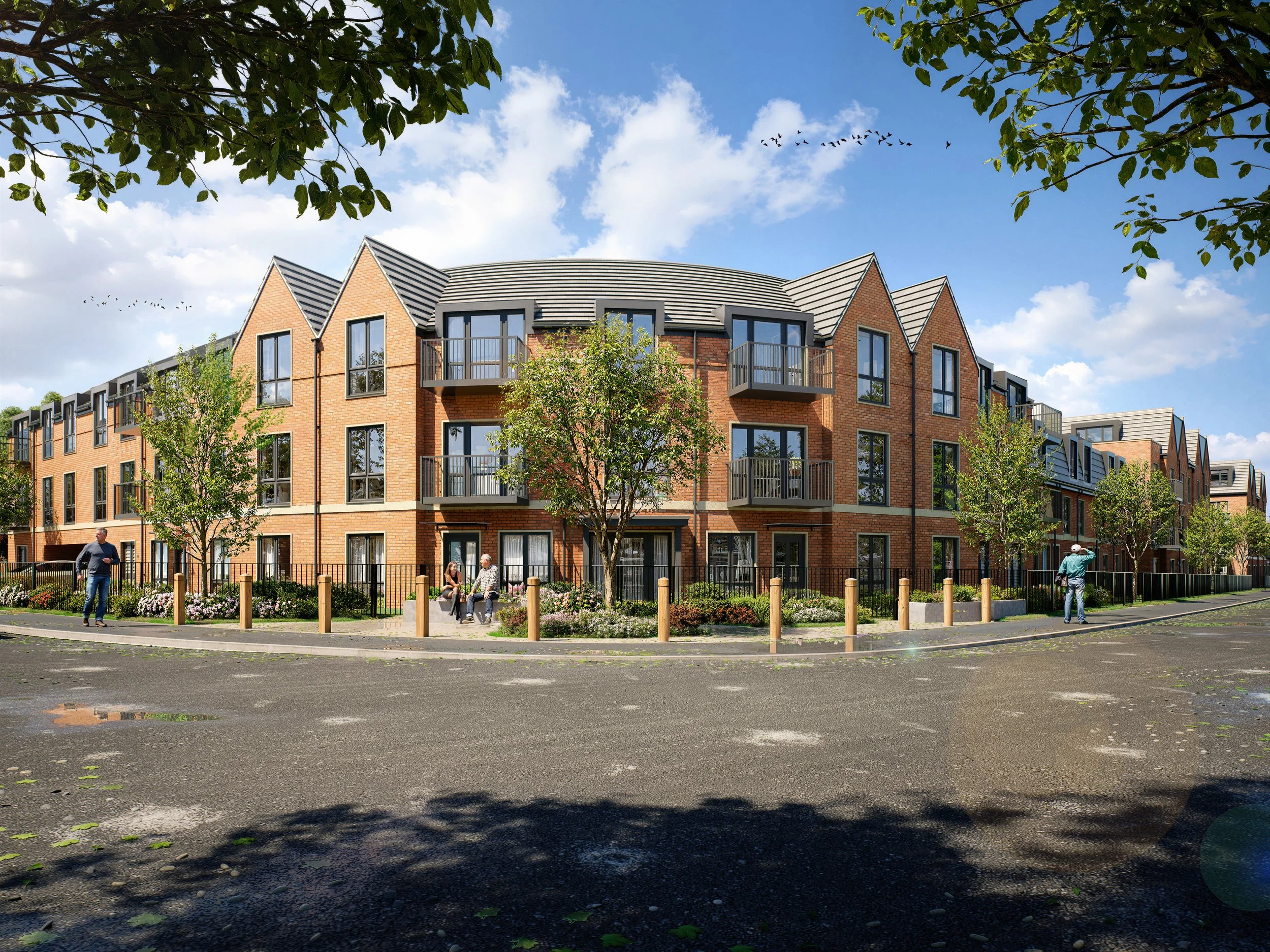 A modern red brick apartment building with multiple balconies, surrounded by greenery and trees, with people walking and sitting outside on a sunny day.