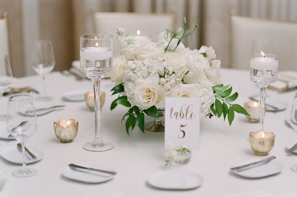 Elegant wedding table with white roses, floating candles, gold votives, and a "Table 5" card on a white tablecloth.  the newbury boston wedding
