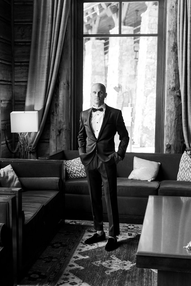 Groom in a classic black tuxedo standing by a large window in a rustic lodge.