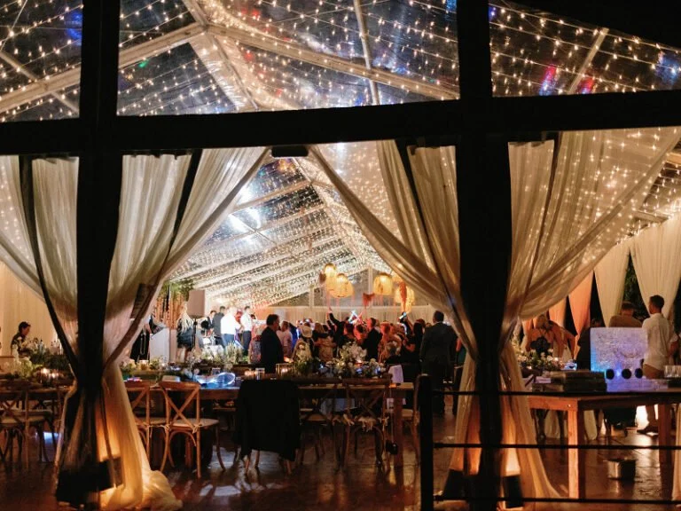 Evening wedding reception under clear-top tent with twinkle lights and draped curtains in mountain setting.