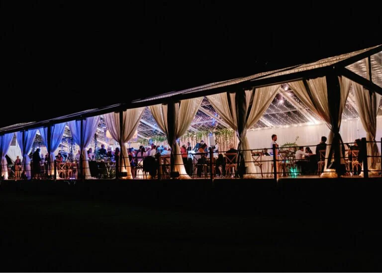 Nighttime wedding reception in clear-top tent glowing with lights, draped in elegant fabric, Colorado wedding design.
