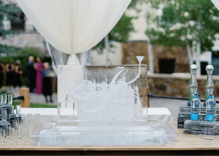 Elegant ice luge sculpture at wedding reception, shaped for pouring drinks, with draped white backdrop.