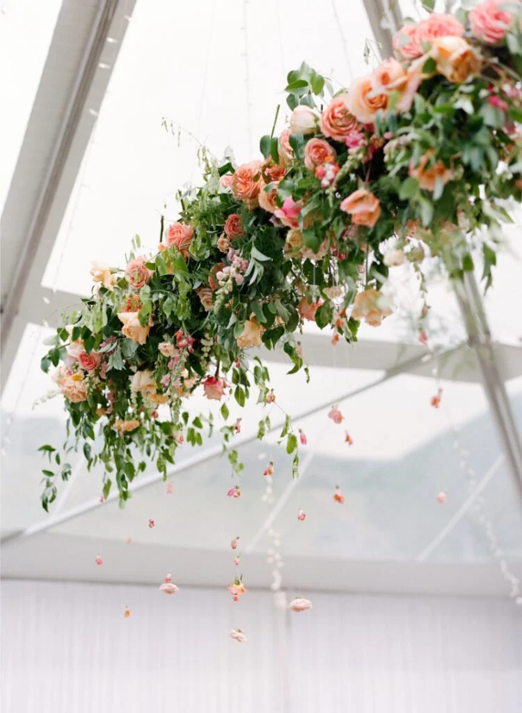 Overhead floral and drapery installations featuring lush greenery and peach-toned blooms suspended in a tent.