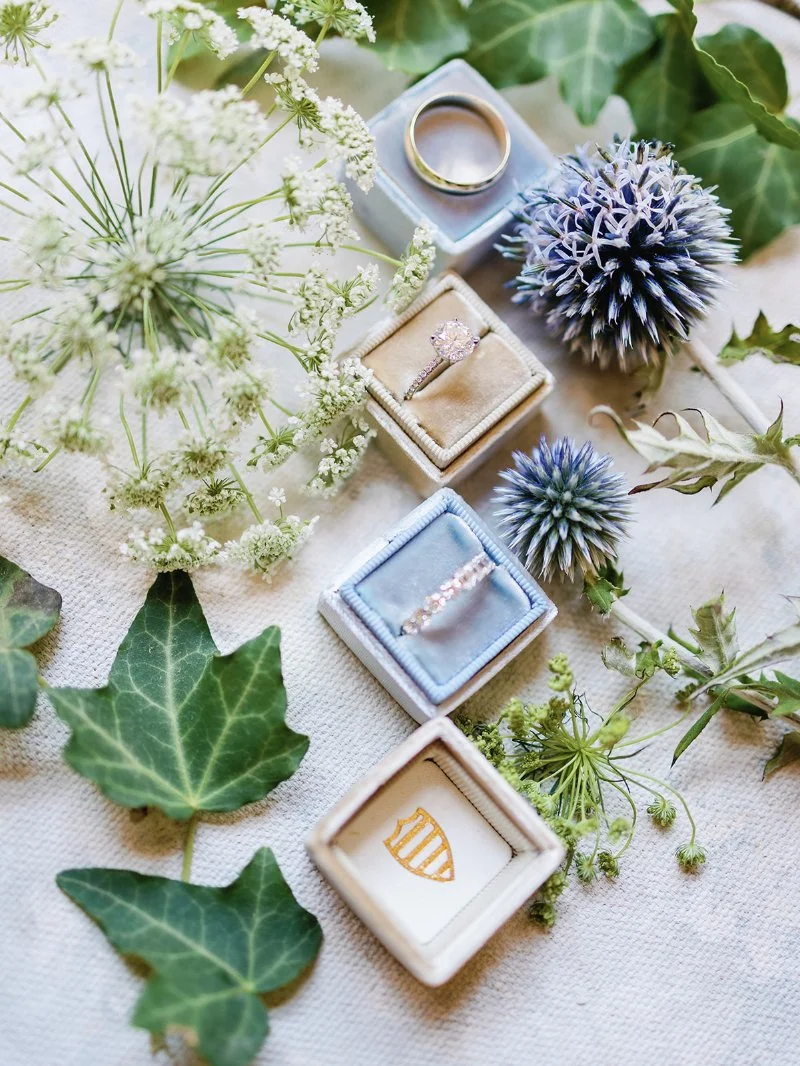 Diamond engagement ring and wedding band in velvet boxes surrounded by delicate white and blue flowers.