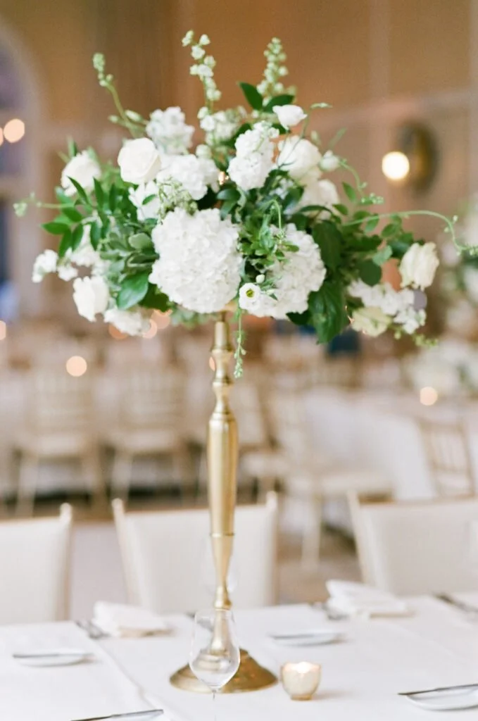 Elegant white floral centerpiece with hydrangeas and roses in a gold vase at a Wedding at The Mount, set in a classic reception space with ivory chairs and soft candlelight.