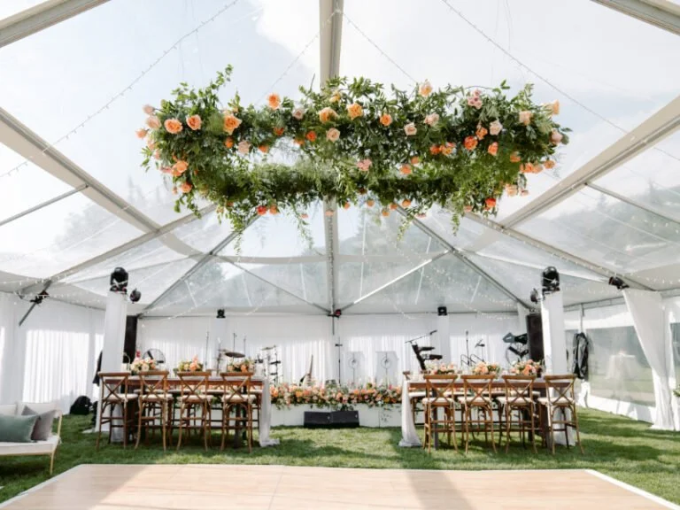 Lush floral chandelier suspended in a clear-top tent above a reception setup with wood chairs.