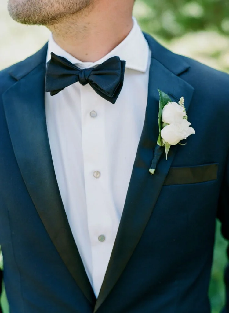 Close-up of a groom in a navy tuxedo with a black bow tie and white boutonniere, captured during a wedding at The Cliff House.