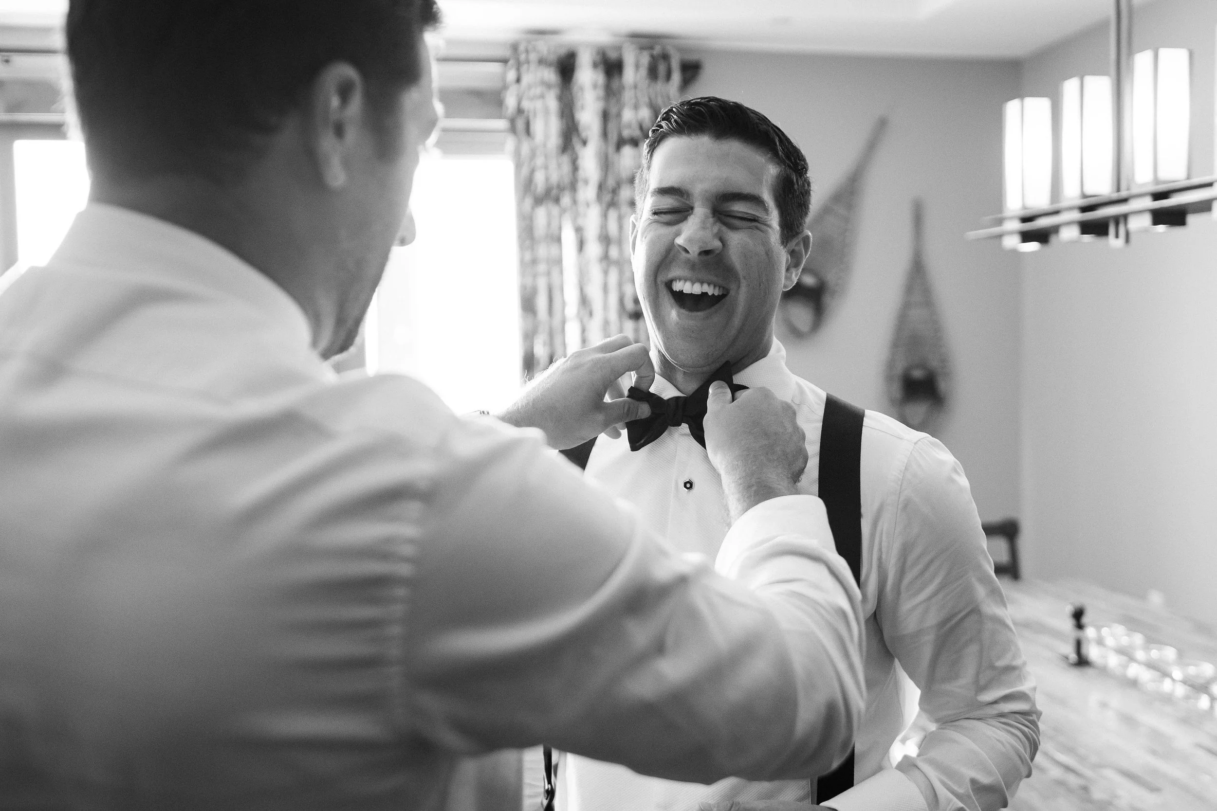 a groom laughing at his friend helping him put on his tie before his wedding ceremony
