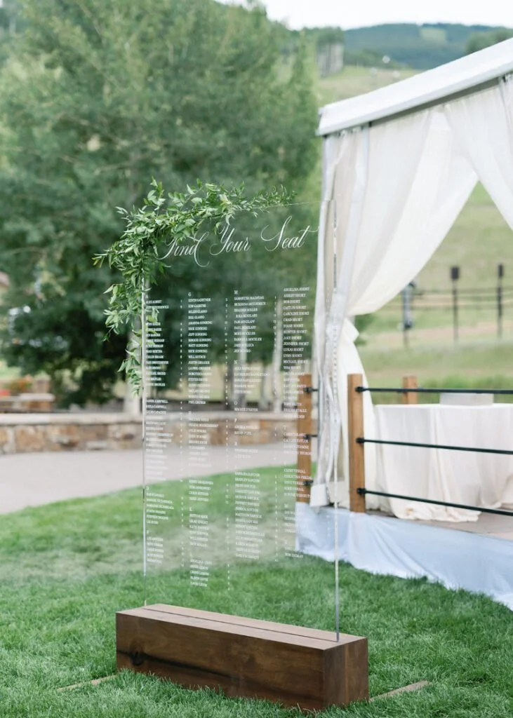 Acrylic seating chart with greenery and wood base, offering elegant and modern wedding design ideas.
