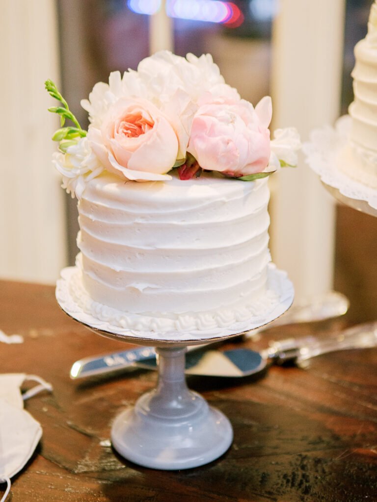 Small white wedding cake with textured buttercream frosting, topped with blush roses and peonies, displayed on a pale pedestal stand.