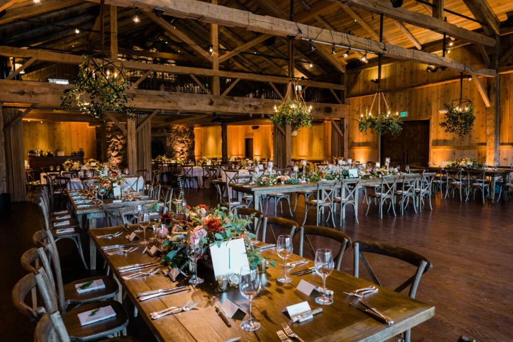 Wooden beams, long farm tables with colorful floral centerpieces, hanging greenery chandeliers, and warm golden lighting create a cozy wedding atmosphere.