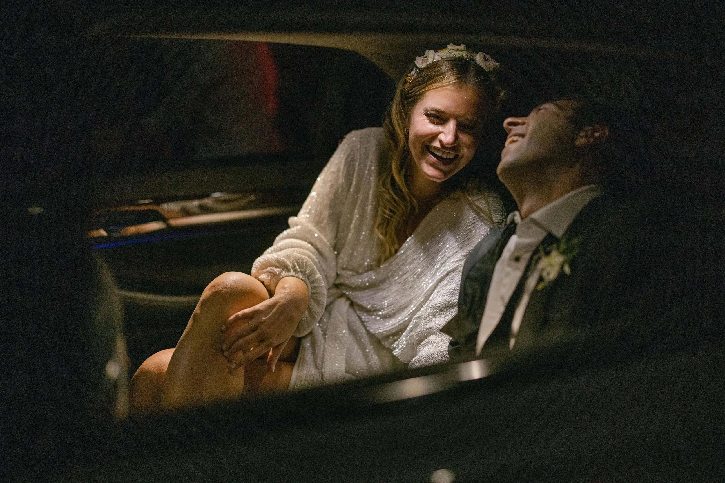 a happy bride and groom laughing together in a car before they leave the venue