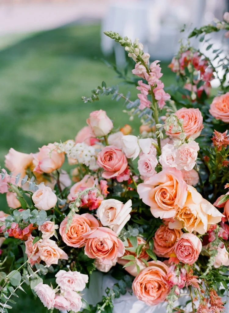 Close-up of a lush floral arrangement featuring peach, blush, and coral roses mixed with greenery and delicate pink snapdragons, set outdoors on a sunny day.