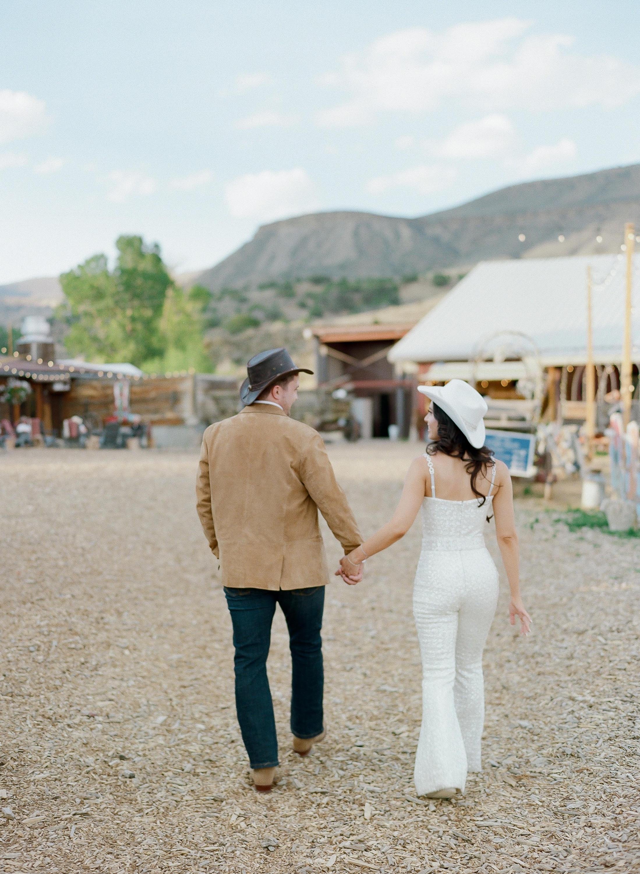 a bride and groom holding hands walking away wearing cowboy hats