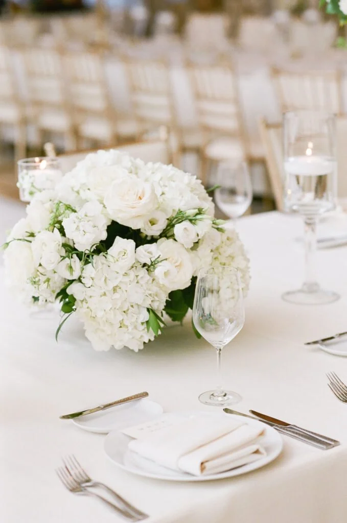 Elegant white floral centerpiece with roses and hydrangeas on a crisp reception table setting, captured during a wedding at The Cliff House.