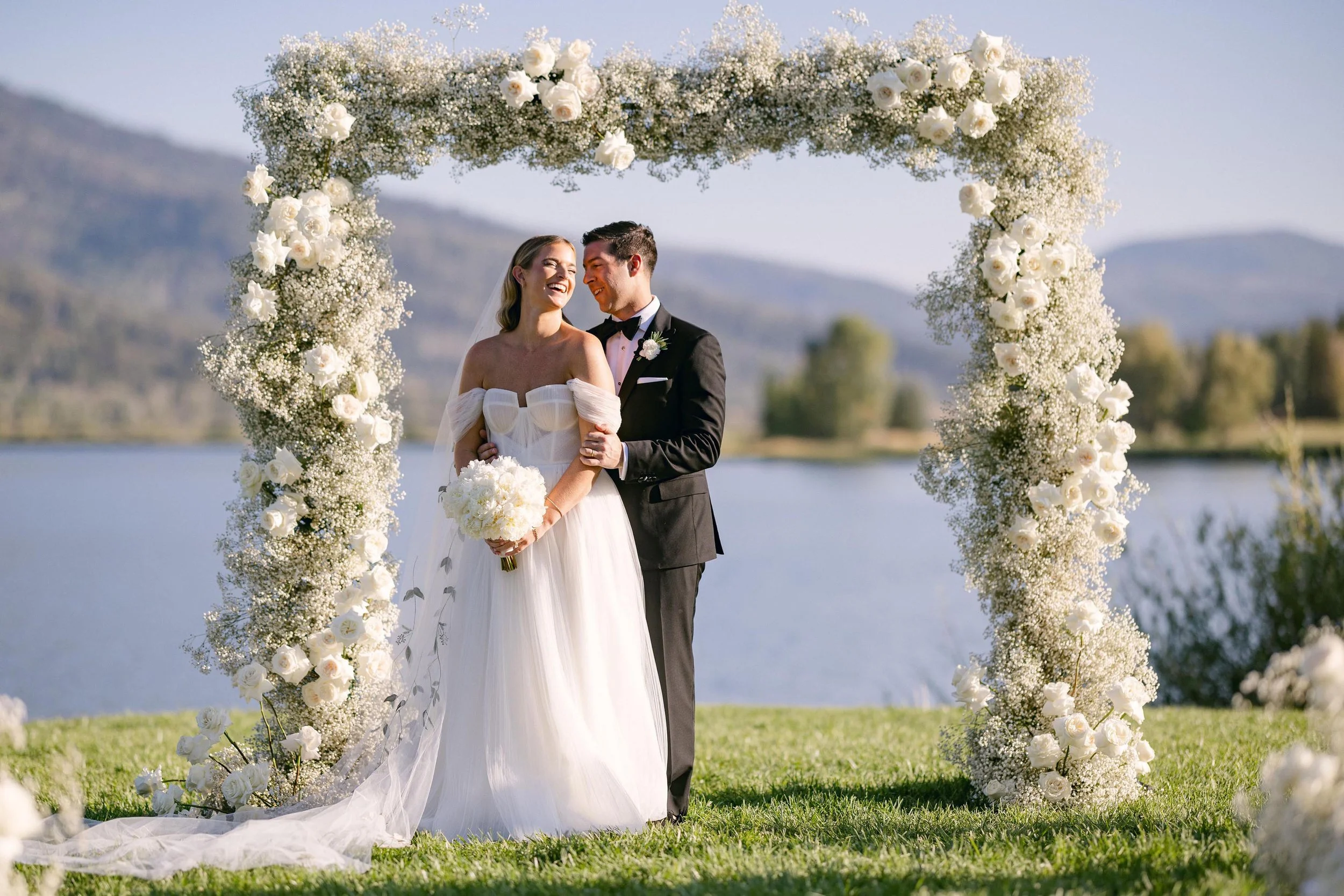 a bride and groom laughing with each other under a white floral arch in front of lake in colorado