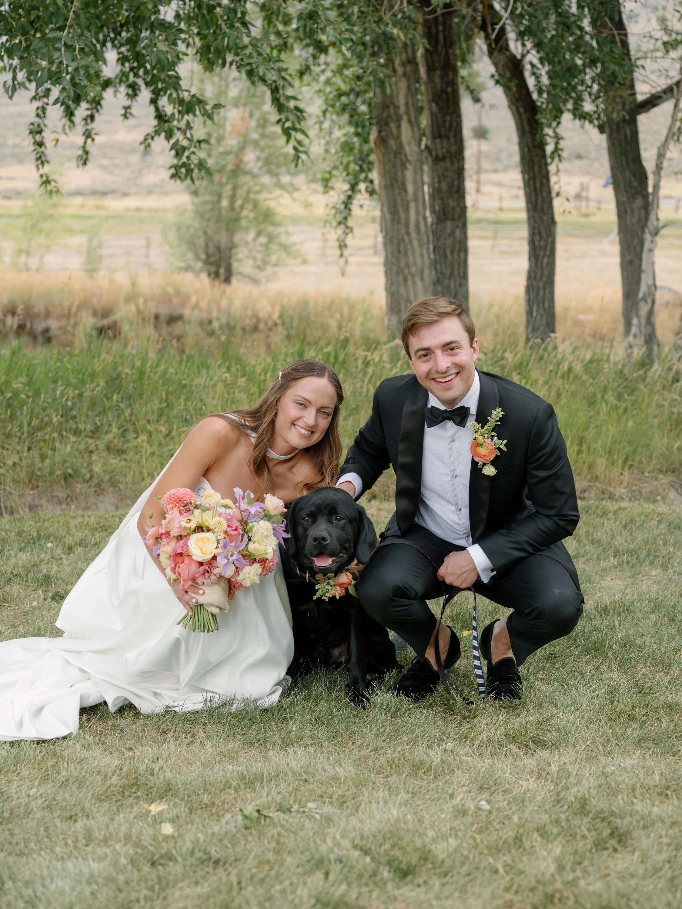 a bride and groom posing with their black labrador on a field in colorado