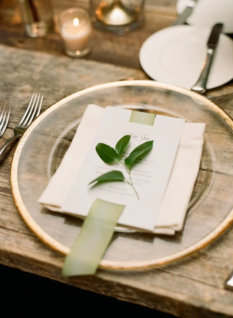 Elegant wedding menu placed on folded napkin with green ribbon and fresh leaf accent on glass plate.