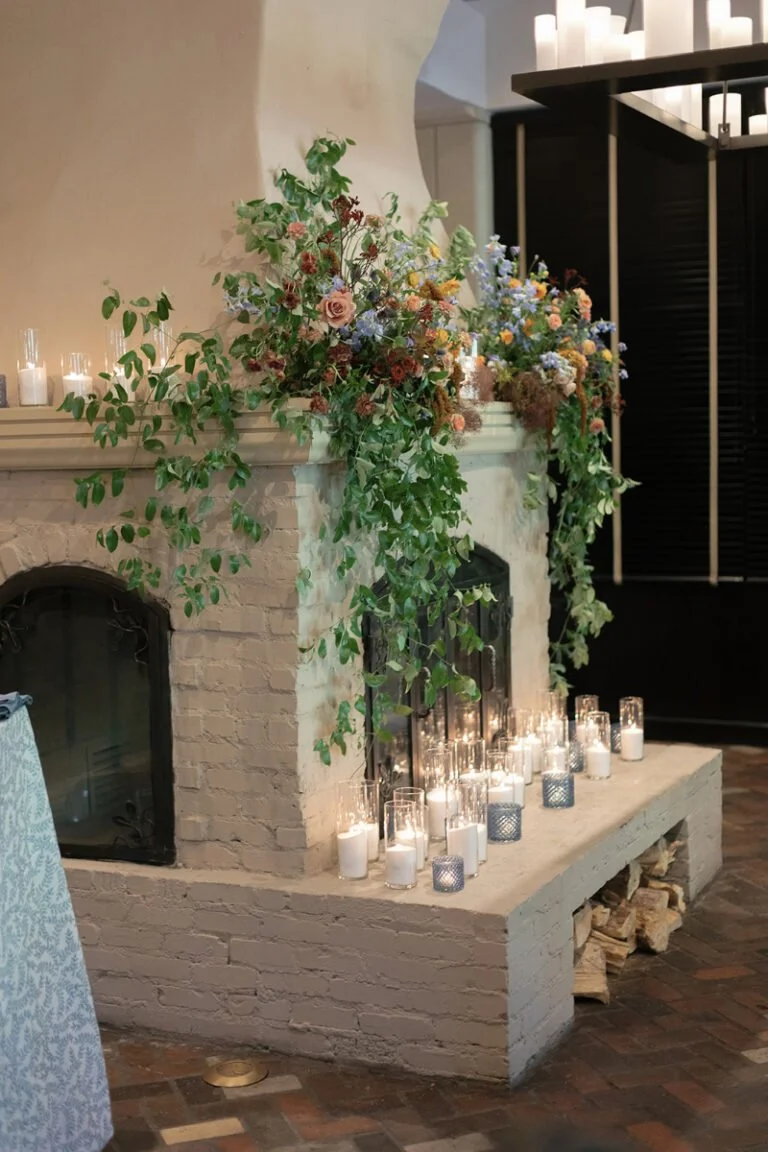 Romantic fireplace floral installation styled by wedding planner with candles, greenery, and soft garden blooms.