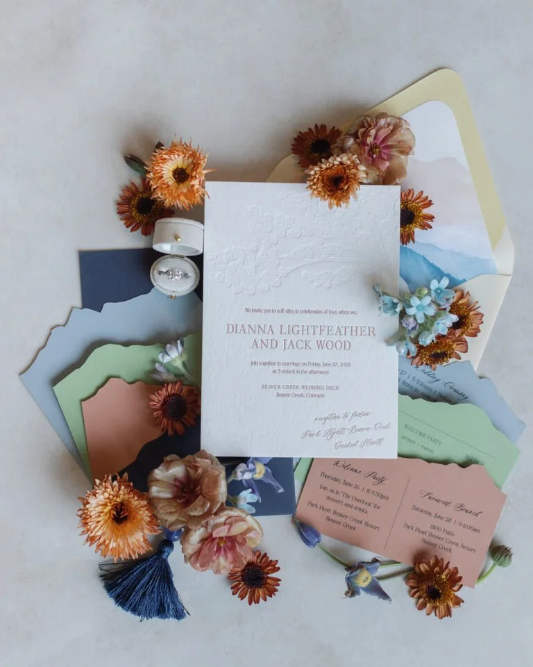 Elegant embossed invitation with colorful envelopes, orange and pink flowers, a diamond ring, and tassels arranged artfully on a neutral background.