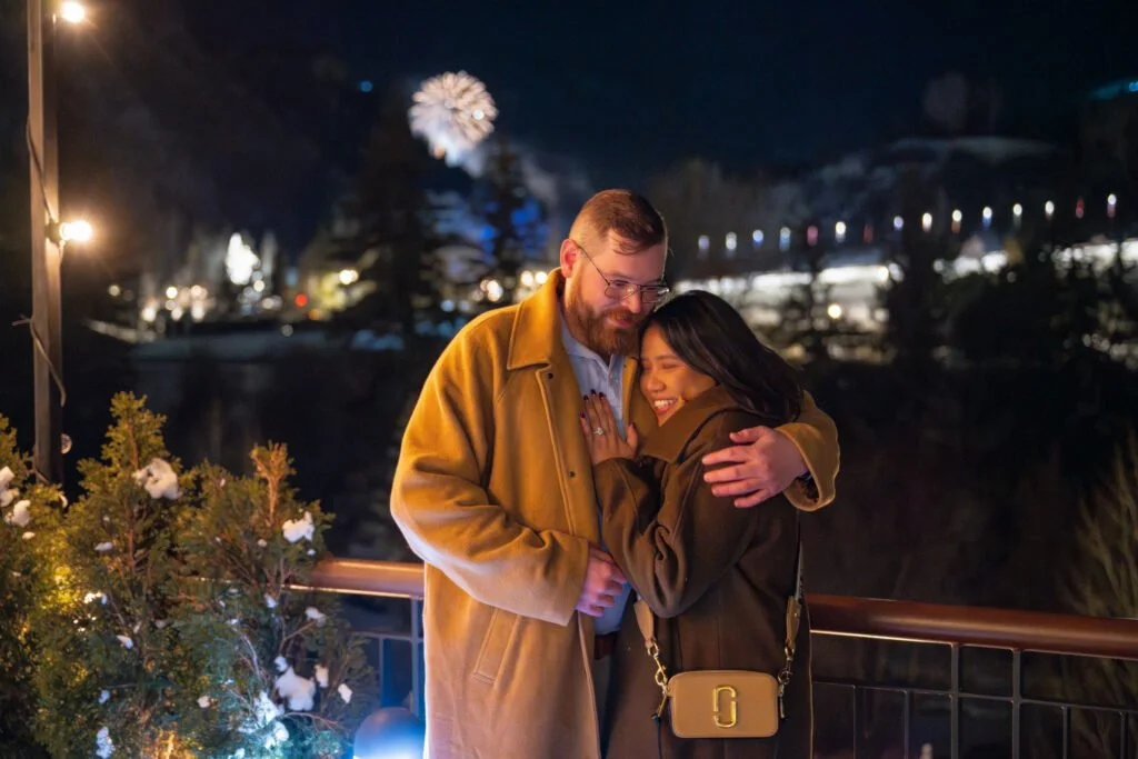 Engaged couple shares an intimate winter moment under fireworks on a snowy balcony at night.