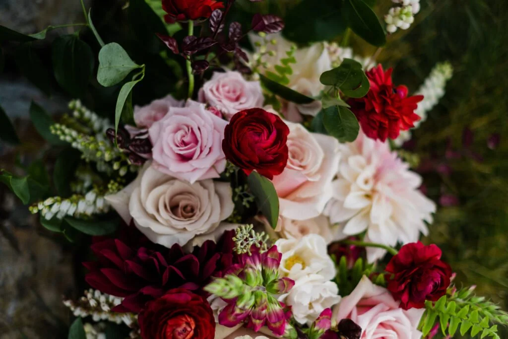 Close-up of a vibrant floral arrangement featuring deep red ranunculus, blush and mauve roses, white dahlias, and lush greenery.