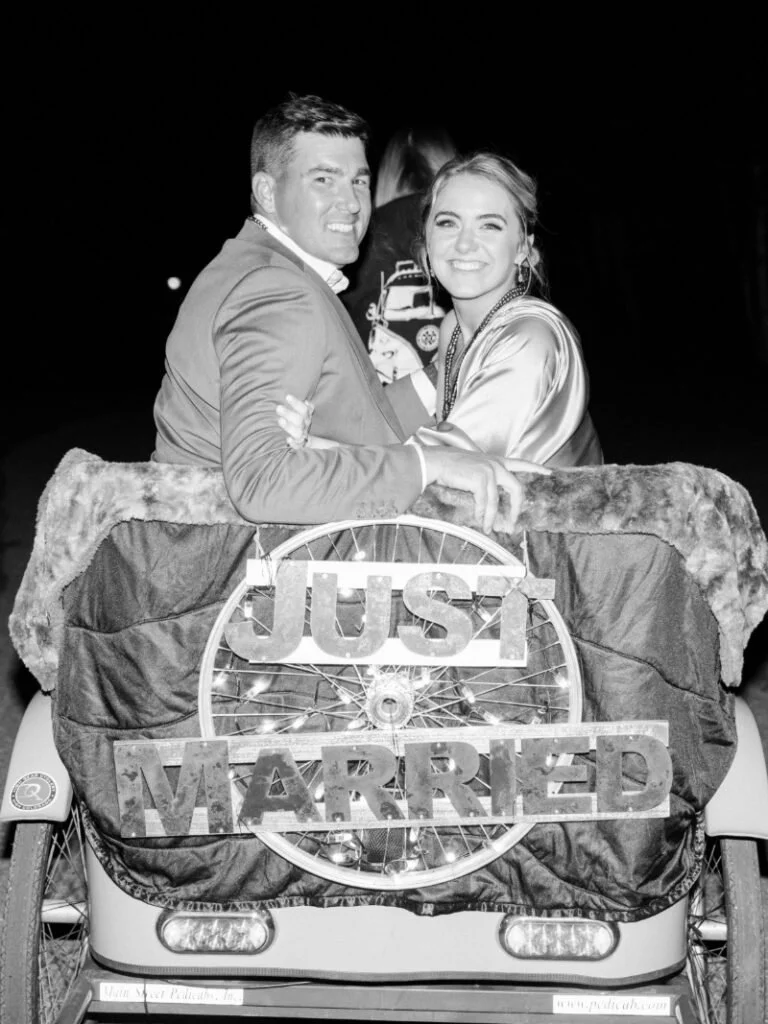 Bride and groom smiling in vintage getaway car during evening wedding sendoff.