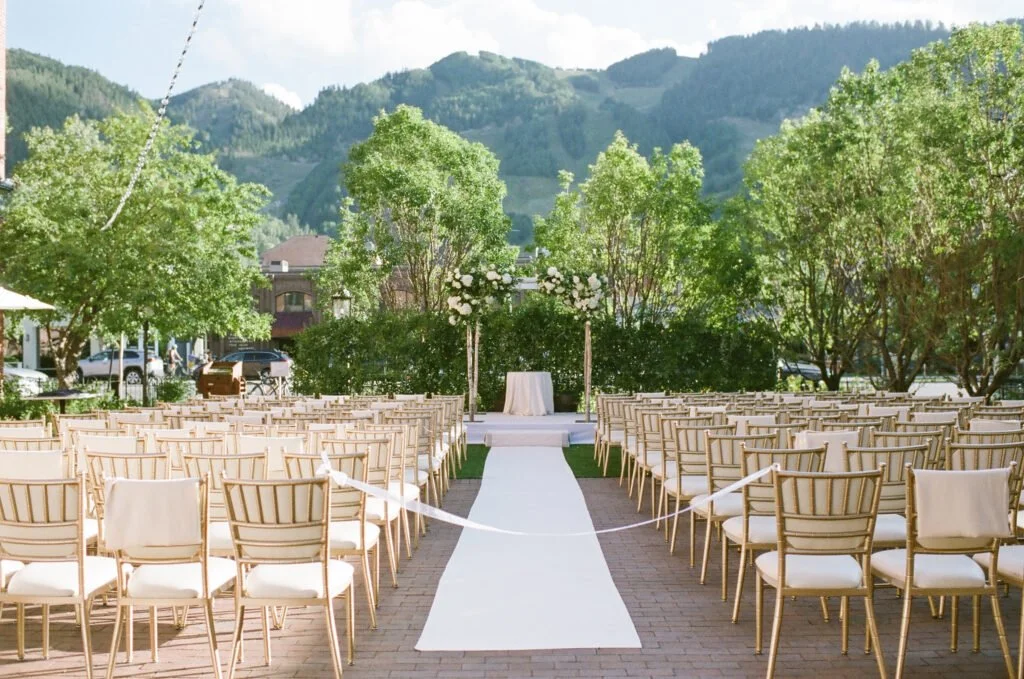 White aisle runner lined with gold chairs leading to a floral arch, set against a lush green mountain backdrop on a sunny day.