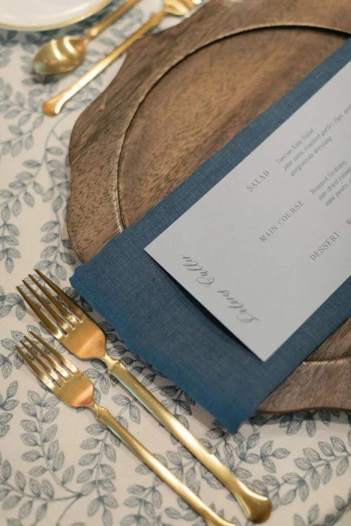 Elegant 2026 wedding design trend featuring layered place setting with blue linen, wood charger, and gold flatware.