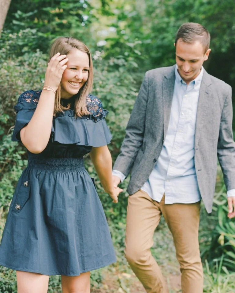 Smiling couple walks hand in hand through a wooded path during a lighthearted outdoor engagement session.